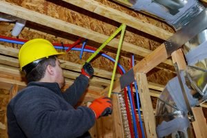 Male builder measuring plastic pipes with new home construction in hot and cold blue and red pex pipe layout in pipes exposed beams
